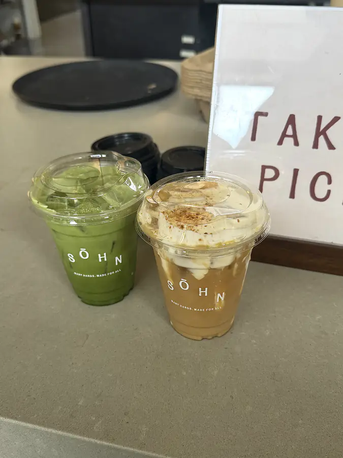 Two iced drinks in branded cups on a counter beside a hand-lettered sign, in warm afternoon light.
