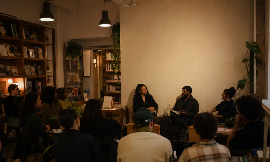 An audience seated close together at a small evening event inside a bookshop, warm lamplight.
