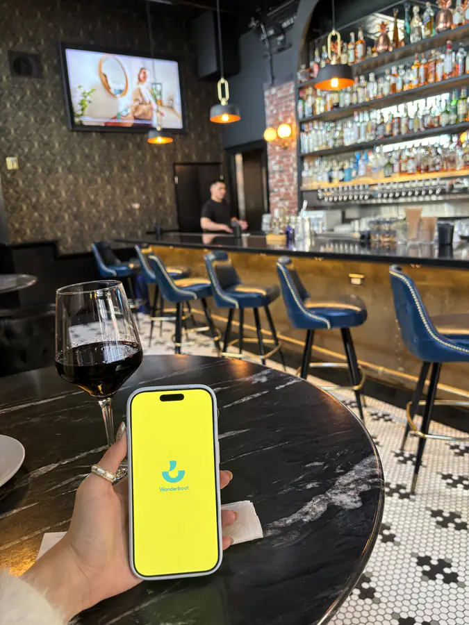 A hand holding a phone with the Wanderboat app open, on a marble bar counter in front of blue bar stools.