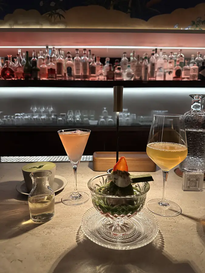 Two cocktails on a counter in front of a backlit shelf of bottles at night.